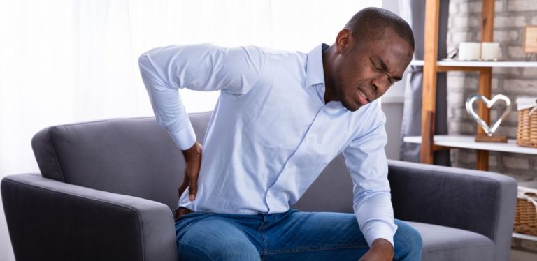 A man sitting in a chair with a pained look on his face while placing his hand against his lower back.