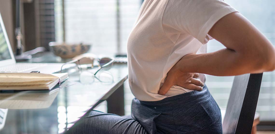 A woman sitting at a desk in a chair holding her lower back.