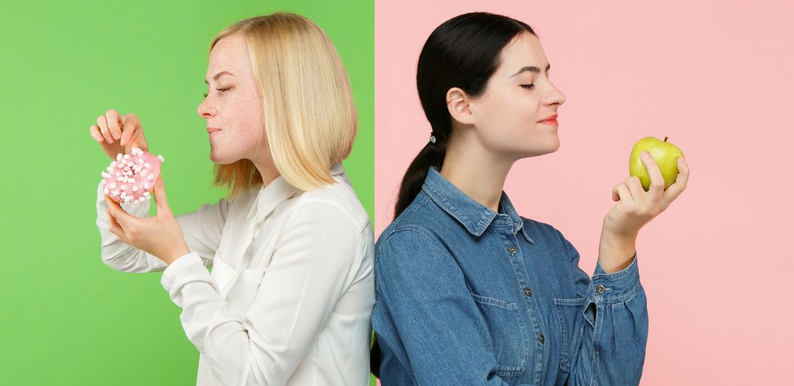 Two women standing back to back: one is holding a doughnut and the other is holding an apple.