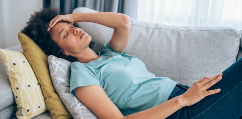 A Black woman laying on a couch with her hand on her forehead.