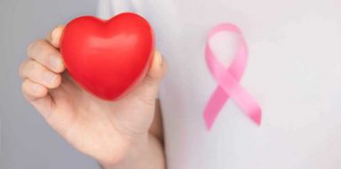 A man wearing a pink shirt, a pink ribbon pin, and holding a red foam heart.