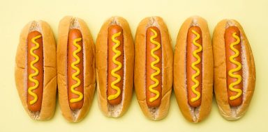 Six hot dogs in buns with mustard on top, on a yellow background.