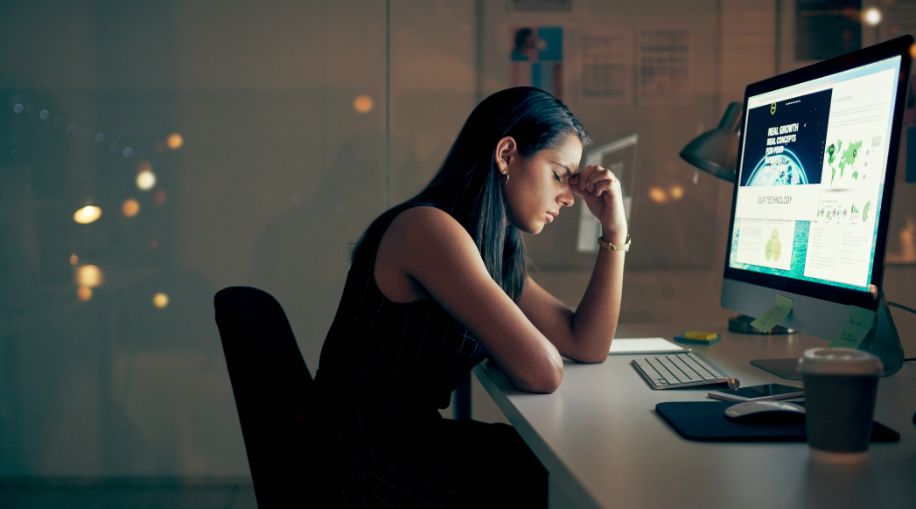 A woman sitting in front of a computer, holding her fingers up to her forehead.